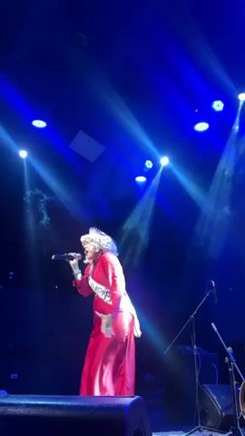 A person wearing a red dress and a sash is singing into a microphone on a stage. The stage is lit by bright blue spotlights, and smoke can be seen in the air. There are microphones and other stage equipment visible in the background.