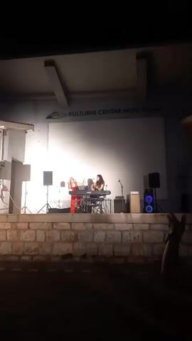 Two women are performing on an outdoor stage at night. One woman, wearing a red outfit, stands to the left and appears to be singing. The other woman, wearing dark clothing, sits behind a keyboard and plays an instrument. The stage is made of stone, and there are speakers and lighting equipment. Behind the stage, there is a building with a large white screen and text that reads "KULTURNI CENTAR NOVI PAZAR". The audience is visible in the foreground, with people seated and facing the stage. The overall atmosphere suggests an evening concert or performance.