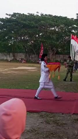 A person in a white uniform, possibly a student or cadet, walks across a red carpet outdoors. They are carrying a small, flat, orange and yellow object. Behind them, two Indonesian flags, red and white, are displayed on poles. To the right of the flags, a large floral arrangement is visible. In the background, trees and a low wall are present, and several people, including some in military camouflage, are standing. The sky is overcast.