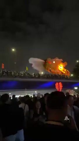 At night, a large illuminated golden dragon sculpture breathes a stream of water over a bridge filled with people, with a crowd of spectators watching from below. The bridge is lit with blue lights, and there are several flags along its railing. The dragon, with red and yellow lights in its eyes, is positioned against a dark sky with scattered clouds. The crowd below includes a mix of men and women, some holding phones to capture the spectacle. Buildings and streetlights can be seen in the background.