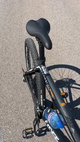 The video shows a first-person view from a bicycle. The bike is black with orange accents and has a water bottle attached to the frame. The road ahead is paved and appears to be a country road with grassy verges. In the distance, there is a valley with trees and a city or town. Mountains can be seen in the far distance under a clear blue sky. The camera pans slightly, showing the road winding and sloping downwards. The overall impression is one of a scenic bike ride on a sunny day.