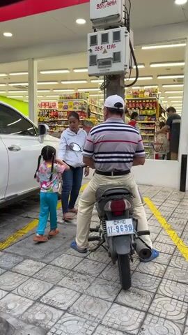 A woman and a young girl are standing next to a white car. The woman is holding a motorcycle handle. A man wearing a striped shirt and a white cap is sitting on the motorcycle. He is wearing light-colored pants and blue shoes. The girl is wearing a colorful shirt and blue pants. They appear to be interacting. Behind them is a store with shelves stocked with products. There is a utility box mounted on a pole in the background. The ground is paved with square tiles.