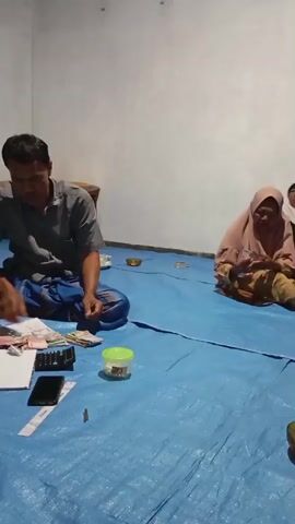 A man and two women are seated on a blue tarp spread across a floor. The man, on the left, wears a gray shirt and a sarong. He is looking down at a piece of paper and appears to be counting money, with a calculator and stacks of Indonesian Rupiah banknotes on the tarp beside him. A green container and a small object are also on the tarp.

To the right of the man, a woman wearing a light pink hijab and patterned clothing is seated and also appears to be handling money or papers. Behind her, another woman in a white hijab is partially visible. Further to the right, a woman wearing a black hijab and a white top is seated on a striped mat, facing the other two individuals and appears to be talking.

The environment suggests an indoor setting, possibly a home, with a plain wall in the background and a doorway with a curtain visible on the right. A small bowl containing nuts or snacks is on the blue tarp in the foreground.