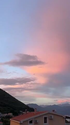 This is a vertical video recording of a sunset over Baošići, Montenegro. The sky is filled with vibrant hues of pink, orange, and purple, with some scattered clouds tinged by the setting sun. In the foreground, the tiled roofs of houses are visible, along with their white walls and air conditioning units. The houses appear to be part of a residential area on a hillside. In the background, a range of mountains stretches across the horizon, some of them silhouetted against the colorful sky. A body of water, likely the Adriatic Sea, is visible between the mountains and the houses. The time of day is clearly sunset, and the weather appears to be clear with some clouds contributing to the dramatic sky. The overall mood is peaceful and serene, capturing the beauty of a coastal sunset. There is no visible text in the video.