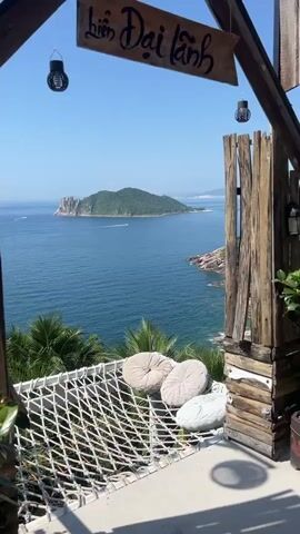 A vertical shot shows a scenic view of the ocean, an island, and palm trees. In the foreground, a white net hammock with three round pillows is visible. Behind the hammock, a rustic wooden structure stands to the right. The ocean is a deep blue, with a green, mountainous island in the distance. The sky is clear and bright blue.