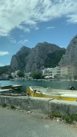 The video captures a scenic view of a harbor or waterfront. In the foreground, there is a concrete barrier and the top of a white boat with a registration number visible. Beyond the barrier, several boats are docked. In the middle ground, the water reflects the blue sky and white clouds. On the opposite side of the water, there are buildings that appear to be apartments or hotels, nestled against a backdrop of lush green hills. Towering over the landscape are imposing, rugged gray mountains with a rough, textured surface. The sky is bright blue with scattered white clouds.