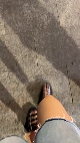 The video shows a first-person perspective of someone walking on a textured sidewalk. The camera is focused on the person's legs and feet, which are wearing denim shorts and black sandals with straps. The ground is made of large, rectangular stone tiles. Shadows of people are visible on the ground, suggesting that the person is walking outdoors in daylight or under artificial lights that cast shadows. The movement of the camera indicates the person is walking forward.