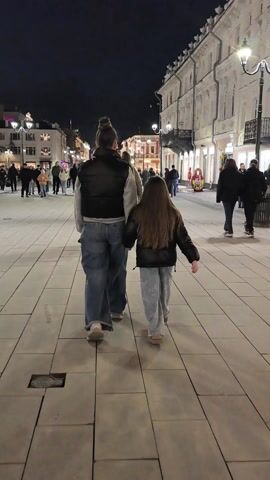 The video shows a woman and a girl walking hand-in-hand down a wide pedestrian walkway at night. The woman is wearing jeans, a light-colored sweatshirt, and a dark vest. Her hair is pulled back in a bun. The girl has long brown hair and is wearing jeans and a dark jacket. The walkway is paved with large rectangular tiles and is well-lit by street lamps. Other people are walking in the same direction, and buildings line the sides of the walkway. The atmosphere appears to be calm and casual.