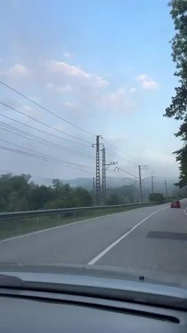 The video shows a view from inside a car driving on a road. On the left side of the road, there is a guardrail and trees. On the right side of the road, there are more trees. There are telephone poles and wires running along the side of the road. The sky is blue with some clouds. The road appears to be wet, suggesting it may have recently rained. In the distance, there are rolling hills covered in mist. A red car is visible in the distance on the right side of the road.