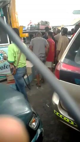 The video shows a gas station or petrol station with several people gathered around the pumps. There are cars parked nearby, including a green car in the foreground. The people appear to be customers waiting to get fuel. A person in a green shirt, presumably a gas station attendant, is visible near the pump. Some of the cars seem to be loaded with luggage, suggesting they may be on a trip. The general atmosphere appears busy, possibly due to a high demand for fuel.