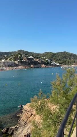 This media file captures a vibrant, sun-drenched beach scene in Tossa de Mar, Spain. The clear, turquoise water of the Mediterranean Sea gently laps against a sandy shore, dotted with numerous beachgoers. The sun is high in the sky, indicating it is midday on a bright, clear day with excellent weather.

The beach is a hive of activity. People are swimming in the shallow, clear water, some closer to the rocky outcrops in the foreground, while others venture further out. On the sand, many individuals are relaxing under colorful beach umbrellas, sunbathing, or strolling along the shore. The shoreline is densely packed with people, suggesting it's a popular time and location for beach tourism.

In the background, a charming town rises up the hills, featuring white buildings with terracotta roofs, typical of Mediterranean architecture. The town blends seamlessly into the lush green, hilly landscape that surrounds the bay. Boats are visible anchored in the water, adding to the scenic coastal view.

In the immediate foreground, there are rocky areas with sparse vegetation, and a few people are sitting on the rocks, enjoying the view. A black railing runs along the bottom right of the frame, suggesting the viewpoint is from a slightly elevated terrace or walkway overlooking the beach. The overall atmosphere is one of leisure, enjoyment, and a typical summer holiday experience. There is no visible text in the media file.