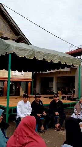 The video shows a group of people gathered outdoors under a green and black canopy. Three men are seated on blue chairs, facing the camera, while others sit in a semi-circle around them. The men are wearing hats, and the person in the center has a "Marlboro" hat. Some of the other individuals are wearing hijabs. In the background, there is a raised platform with a wooden frame, and behind that, a building with a sign that reads "MAHASISWA UNIVERSITAS ISLAM NEGERI." A group of people can be seen sitting at a table in the background of the scene. A person is seen holding a camera, capturing the scene. The setting appears to be a casual outdoor gathering, possibly related to a community event or meeting.