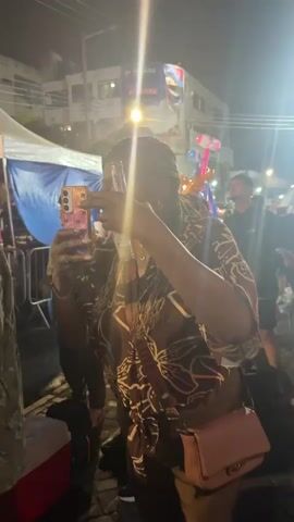 A woman with braided hair is holding a cell phone in her left hand, capturing a video. She wears a patterned brown shirt and a pink crossbody bag. She is outdoors at night with a crowd of people in the background. Some people are holding drinks and looking at the camera or away from it. There are tents and buildings visible, along with streetlights and decorative balloons. The scene appears to be a lively outdoor event.
