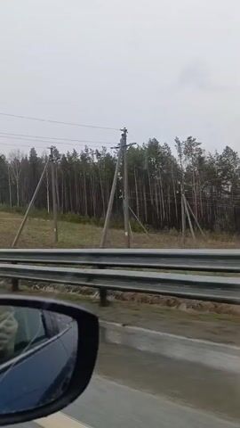 The video shows a view from a car traveling on a highway. The road is wet, and the weather is overcast. A metal guardrail runs alongside the road. Beyond the guardrail, there is a grassy area with trees in the background. Utility poles are visible along the roadside. The side mirror of the car is visible in the foreground, reflecting the interior of the car and the person holding the recording device.