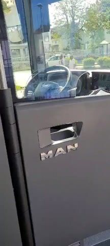 The video shows the interior of a MAN bus. The camera pans from the interior door handle to the steering wheel and dashboard. Outside the bus, buildings, trees, and a street can be seen. A person is visible through the windshield, standing near a taxi sign. The video is shot from the perspective of someone inside the bus, likely the driver.