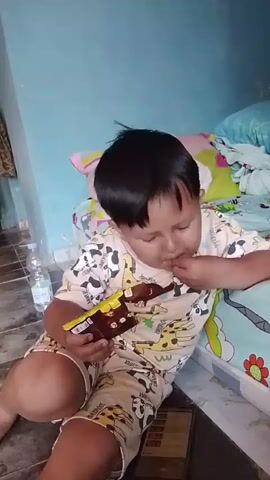 The video shows a young boy sitting on the floor, leaning against a bed. He is holding a chocolate bar and appears to be eating it. The boy is wearing pajamas with a pattern of giraffes and pandas. Behind him, on the bed, are pillows and blankets with a similar pattern. A plastic water bottle is visible on the floor to his left, and a smartphone is on the floor in front of him. The room appears to be a bedroom with light blue walls.