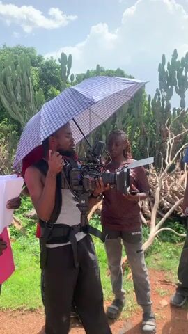 In an outdoor scene set in Farin Yashe, Nigeria, a film crew is in action. The central subject is a man operating a professional video camera mounted on a rig. He wears a gray vest over a sleeveless shirt and dark pants, with a waist support system. To his right, an umbrella with a checkered pattern is held aloft, likely to shield him and his equipment from the sun.

Behind the cameraman, a woman in a burgundy t-shirt and gray pants is also observing the scene, holding a phone in her left hand. To the cameraman's left, another man in a red t-shirt stands close, and further left, a person in a bright pink t-shirt holds a stack of white papers, possibly a script.

The background is lush with vegetation, featuring tall, spiky plants reminiscent of cacti, and dense green foliage. Some dry, gnarled branches are visible, adding to the natural, rustic environment. The ground appears to be a mix of dirt and gravel.

The sky is partly cloudy, suggesting a bright, possibly warm day. The overall atmosphere appears focused and professional, with the crew engaged in their task of filming. The presence of scripts and professional camera equipment indicates a film or video production is underway. No visible text is present on the clothing or equipment, aside from what might be a logo on the woman's t-shirt, which is not legible.