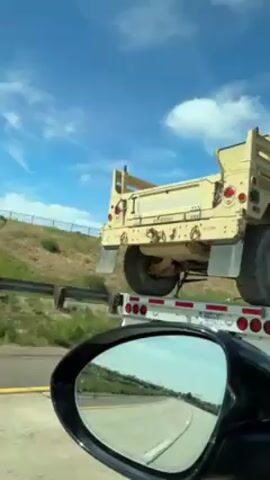 A vehicle films from the driver's side on a sunny day, capturing a flatbed trailer transporting a tan Humvee. The Humvee lacks doors, with an open driver's side, and appears to be missing the front section of its roof. The flatbed is secured with chains, with red and white stripes on its side. The Humvee's back end and large tires are visible. The sky is blue with some clouds. The side mirror of the filming vehicle reflects the highway.