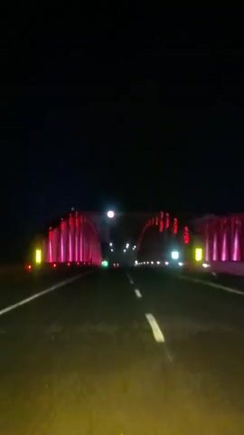 The video shows a first-person perspective of a vehicle driving on a highway at night. The highway is illuminated by the vehicle's headlights and streetlights. The vehicle is approaching a bridge or overpass that is decorated with red and pink lights. The lights are arranged in a pattern along the structure, creating a vibrant and colorful display against the dark sky. As the vehicle gets closer, the structure appears to be a tunnel or underpass, as the lights become more enclosed. Traffic signs are visible, indicating speed limits. The road surface is asphalt, and white lines mark the lanes. The overall atmosphere is one of a modern, well-lit urban or suburban environment at night.