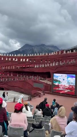 The video shows an outdoor amphitheater with tiered red seating. A crowd of spectators, many wearing red hats, are seated in the foreground, facing a stage. On the stage, performers in traditional attire are participating in a cultural performance. The backdrop features a large mountain range with cloudy skies. A large screen is visible on the right side of the stage displaying images and text, possibly providing information about the performance or location. People are seen standing and walking around the stage area, interacting with each other, and taking photos. The overall atmosphere appears to be one of cultural tourism and entertainment.