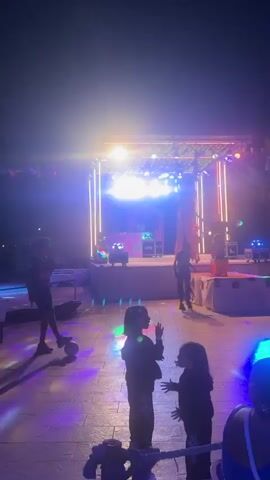 The video shows an outdoor event at night with a stage and a dance floor. There are people of various ages in the scene, including children in the foreground and adults in the background. The stage is lit with bright, colorful lights, and there are speakers and DJ equipment on it. People are dancing and enjoying themselves. Some people are watching the performance from a distance, while others are closer to the stage. There are flags strung across the top of the stage area, and palm trees are visible in the background. The ground appears to be paved. The lighting creates a festive atmosphere.