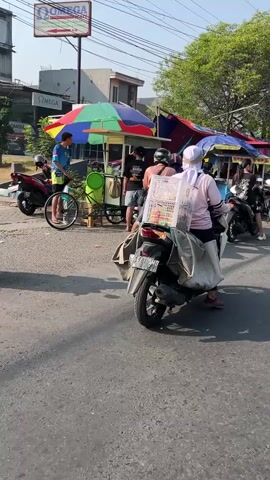 The video shows a street scene with several people and vehicles. In the foreground, a motorcycle is parked on the right side of the road. It is loaded with goods covered by a tarp, and a person wearing a white head covering and a pink shirt is seated on it. Behind the motorcycle, there are food carts with colorful umbrellas. One cart appears to be selling drinks from a green container. Several people are gathered around the carts, some appear to be customers or vendors. A person in a blue shirt is standing near a cart, holding what looks like a bicycle wheel. In the background, there are buildings, trees, and power lines, suggesting an urban or semi-urban environment. The lighting indicates it is daytime, likely morning or afternoon. Several other motorcycles are visible parked or moving in the background.
