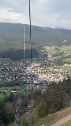 A high-angle drone shot shows a mountain village from a ski lift. The village is nestled in a valley surrounded by forested mountains. The ski lift runs vertically down the frame, with the camera looking down and to the right. Several red ski gondolas are visible on the lift line, and the village is densely packed with buildings of various sizes and colors. The mountains are covered in green trees, and the sky is cloudy. The scene appears to be a scenic, natural landscape with a human settlement.