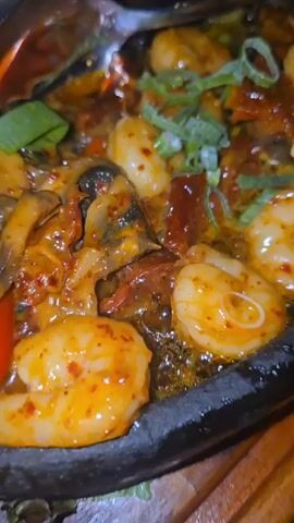 The camera captures a delicious, sizzling shrimp dish from a high angle. The shrimp are arranged in a black, oval-shaped skillet, filled with a rich, reddish-orange sauce. Chopped bell peppers and mushrooms are scattered throughout, and green onions garnish the top. The skillet rests on a wooden serving board, revealing a vibrant, cooked shrimp with spices on the surface. The background shows a blurred view of a silver bucket and a clear bottle, likely indicating a restaurant setting.