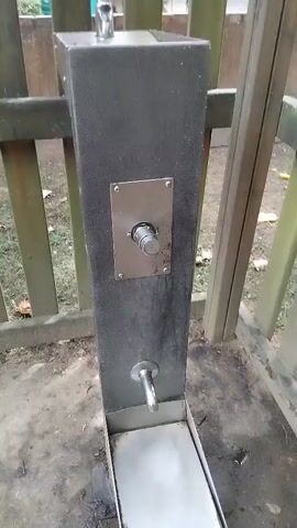 The video shows a close-up of a metal outdoor water dispenser. The dispenser is attached to a dark gray rectangular post, which is mounted on a concrete or dirt ground. A wooden fence is visible in the background. The dispenser has a silver plate with a button or dial on it, and a metal spout below. A hand enters the frame and turns the dial. The dial appears to have some markings, but they are not clear enough to read. The dispenser is likely in a public outdoor space, perhaps a park or a trail, given its design and placement. The lighting suggests it is daytime, and there are no obvious signs of extreme weather. The overall impression is of a functional, utilitarian object in a natural or semi-natural setting.