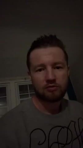A man with short brown hair and a beard is looking at the camera. He is wearing a gray t-shirt. He touches his forehead, then his hair, and then adjusts his shirt. The background is dimly lit with what appear to be windows.