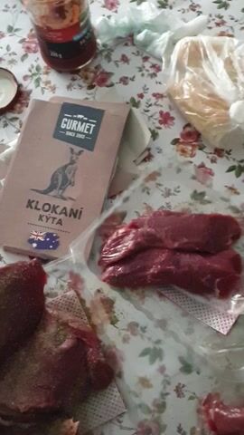 The video opens with a top-down view of a table covered with a floral tablecloth. In the center of the frame, there are two pieces of raw, dark red meat, possibly kangaroo based on the packaging. To the right of these pieces, another piece of raw meat is visible, sprinkled with a light brown powder, possibly a spice or seasoning. Above the meat, there is a clear plastic bag with some light brown powder inside. To the left of the meat is a brown cardboard package with the word "GURMET" and an image of a kangaroo, along with the text "KLOKANI KYTA" and an Australian flag. A glass jar with a red lid and some sauce is also on the left side of the table, along with a small metal lid. A green object, possibly a piece of vegetable, is lying near the bottom left of the frame. In the second and third segments, a hand holding a cylindrical shaker is visible. The hand is shaking the shaker over the meat on the right side of the frame, sprinkling it with a light brown powder. The action is blurry due to the movement of the hand and shaker.
