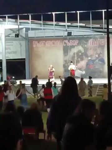 An outdoor evening scene depicts a cultural event with a stage and a large audience. On the brightly lit stage, two individuals in traditional Bulgarian folk costumes are performing. A woman on the left sings into a microphone, wearing a white headscarf, a dark embroidered sleeveless dress, and a white blouse. A man on the right, in a white shirt, red sash, and dark pants, stands beside her. The stage backdrop features Cyrillic text reading "БЪЛГАРСКИ СЪБОР" (BULGARIAN ASSEMBLY/FAIR) and an historical image on the right side. In front of the stage, on a grassy area, approximately 10-15 people are actively participating in a folk dance, forming a circle or line while holding hands. They wear a mix of casual modern clothing and some elements of traditional attire. In the darker foreground, numerous spectators are visible, some sitting on chairs and others standing, watching the performance. Folk music with Bulgarian singing is playing throughout the scene.