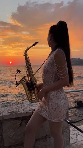 A woman stands by the sea at sunset holding a saxophone. She is wearing a sparkly dress and her hair is in a ponytail. The sun is setting over the water, casting a warm glow on the scene. The sky is filled with colorful clouds. There are mountains in the background. The woman is looking at the camera and smiling. The scene is peaceful and romantic. The time of day is sunset. The weather is clear. The location is by the sea, likely a beach or a promenade, with a stone wall in the foreground. The woman appears happy and confident. There is no visible text in the media file.
