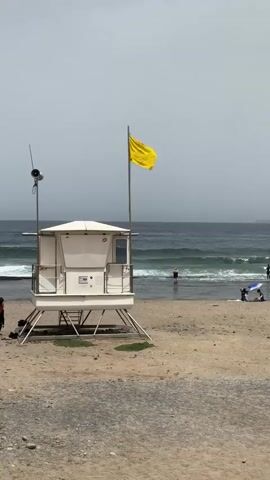 The video depicts a beach scene on a cloudy day, with a lifeguard tower prominently featured in the foreground. The tower is white with a white roof and has a small enclosed cabin at the top. A yellow flag is flying from a pole attached to the tower, indicating potentially hazardous conditions. A speaker or siren is mounted on a pole next to the tower.

The beach itself is sandy and appears to be moderately populated. In the middle ground, the ocean has noticeable waves, suggesting conditions suitable for surfing. Several individuals are visible in the water, some appearing to be surfers or wading. Further down the beach, a couple is sitting under a blue umbrella. A person carrying a surfboard walks on the sand near the left edge of the frame.

The overall atmosphere is calm, despite the yellow flag which typically signals caution. The cloudy sky suggests it might be late morning or early afternoon. The setting is clearly a coastal environment, and the mention of Playa de las Américas provides a specific geographical context. No discernible text is visible on the lifeguard tower or in the surrounding environment.