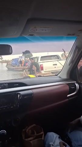 The view is from inside a vehicle, likely a car, looking out the front windshield towards a river or large body of water. A small ferry boat is in the water, carrying at least two vehicles: a white pickup truck and a smaller blue truck. Several people are on the boat, interacting with the vehicles or each other. One man is standing near the white truck, possibly helping with loading or unloading. The boat is close to the vehicle from which the video is being taken. The interior of the car is visible, including the dashboard with a radio and some storage areas. A person's hand is visible in the lower right corner. The sky is overcast.
