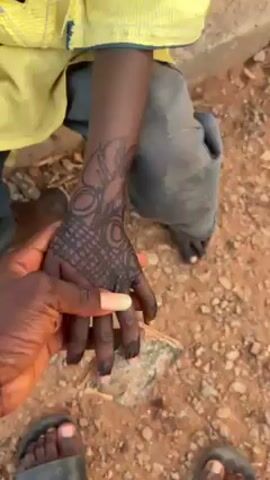A video shows a person’s hand covered in black henna designs, moving over to the bare feet of a child, also adorned with henna. The camera pans down to reveal a small turtle, then focuses back on the feet. The ground is dirt with small rocks. The person filming is wearing sandals.