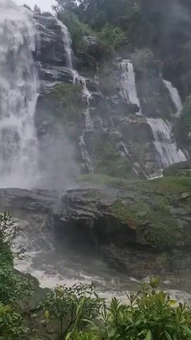 A waterfall cascades down a rocky cliff face surrounded by lush green foliage. The water is white and foamy as it plunges into a pool below. Mist rises from the base of the falls, obscuring the lower parts of the cliff. The sky above is overcast.