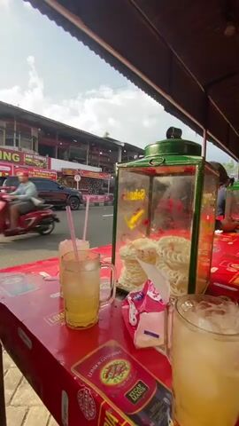 The video is shot from a low angle, showing a red table in the foreground. On the table are two glasses of iced beverage with straws, a glass display case filled with white crackers, and some pink packaging. In the background, a street is visible with traffic, including a black SUV and several motorcycles. Buildings line the street, some with signs advertising food, such as "SATE KAMBING" and "TRUBUS ASLI". The sky is cloudy, and sunlight is present, suggesting it is daytime. The overall scene appears to be an outdoor seating area of a roadside food establishment.