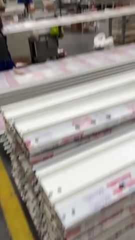 The video depicts the interior of a large factory setting. There are multiple workers, dressed in blue uniforms, actively engaged in various tasks. They appear to be handling long, white or light-colored pieces, possibly components for manufacturing. The factory floor is spacious, featuring metal support beams and overhead cranes. In the foreground are stacks of the white pieces, some covered with a red and white patterned protective layer. There are also cardboard boxes scattered around the area, likely containing supplies or finished products. The overall impression is of a busy manufacturing environment with workers collaborating on an assembly line.