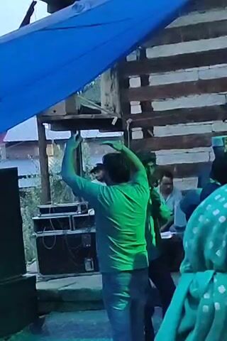 A person in a green t-shirt and jeans dances in front of speakers under a blue tarp. Another person in a light-colored shirt stands next to them. In the background, a person in a white t-shirt appears to be operating DJ equipment, and other individuals are seated. The environment seems to be outdoors, possibly a party or event, with wooden structures visible.