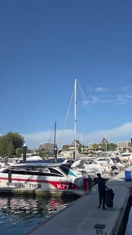 The video begins with a view of a white and red speedboat docked at a marina. The boat has "MOOKSIL TOUR" and "M.77" written on its side. A person in a black shirt and light-colored pants is standing at the stern of the boat. The camera pans to show multiple other boats docked in the marina, as well as palm trees and buildings in the background under a clear blue sky.

The video then shifts to a close-up of the outboard motors of a white boat. The camera moves to show a person walking away from the camera down a wooden dock. They are wearing a dark jacket, a patterned hat, and jeans, and are carrying a black backpack. To the right, another person in a dark shirt and pants is visible.

Next, the camera follows the person walking down the dock, with several boats docked to their right. Palm trees are visible on the left side of the frame, along with buildings and signs, including one that reads "SOLAVITA NOURISH WITH NATURE". The video ends with a view of the water reflecting the sky and buildings.