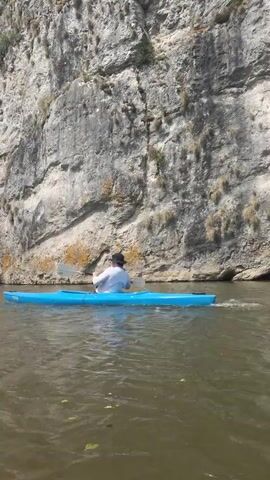 A person is paddling a blue kayak on a river next to a large rock cliff. The person is wearing a white shirt and a hat and is using a paddle to move the kayak forward. The water is murky and there are some leaves floating on the surface. The rock cliff is textured and has some vegetation growing on it. The sky is not visible, but the lighting suggests it is a sunny day.