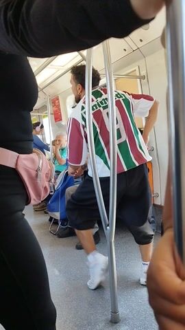 From a slightly elevated perspective, the camera looks down the length of a train car. A man wearing a Fluminense soccer jersey sits with his legs stretched out and partially obstructing the aisle. Other passengers are visible in the background, some seated and some obscured by the camera's positioning. The camera's view is partially obstructed by the handrails, and the arms and chest of an individual holding the camera are visible in the foreground. The overall lighting is bright, and the scene conveys the feeling of a crowded public transportation environment.