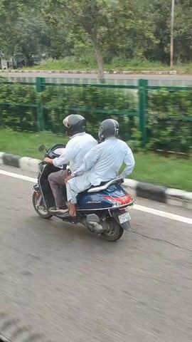 Two men wearing helmets ride on a dark blue scooter. The man in front is driving, and the man behind is seated behind him. They are both wearing light-colored shirts. The scooter is moving on a paved road with a white line to its right. A green fence and trees are visible in the background.