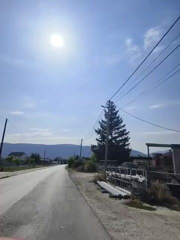 The video shows a road with a car driving down it. The sun is shining and there are trees and houses on the side of the road. There are also power lines running along the side of the road. The video is taken from the perspective of the driver of the car.