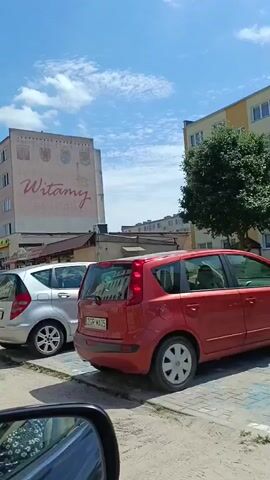 This is a video compilation of several short clips, all filmed from the perspective of someone in a car. The camera pans across a street scene in Gryfino, Poland, on a bright, sunny day. The sky is blue with some scattered clouds.

The primary objects visible are cars parked along the street and buildings lining the road. The buildings appear to be apartment complexes with shops on the ground floor. One building in particular has a large mural on its facade that reads "Witamy" (Welcome in Polish) in cursive red lettering, accompanied by what look like coats of arms. Some of the businesses visible on the ground floor include "LEWIATAN" and a barber shop.

The video captures a variety of vehicles, including a silver hatchback, a black sedan, a blue SUV, a silver van, a green hatchback, and a red compact car. The license plates are visible on some of the cars, indicating Polish registration. The scene appears to be a typical urban or suburban street with parking. There are no people clearly visible in the footage, but the presence of parked cars suggests ongoing activity in the area. The overall mood is peaceful and casual, depicting a normal day in a Polish town.