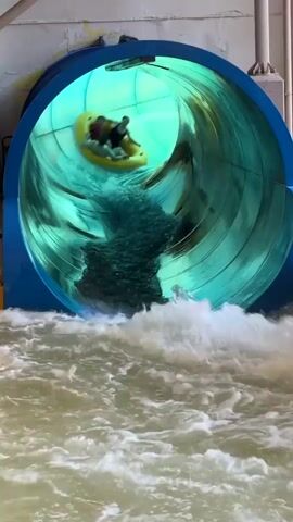 A group of people are riding an inflatable raft down a water slide. The raft is yellow and appears to be carrying at least three people. The water slide is enclosed and lit with green lights, creating a tunnel effect. The raft is exiting the slide into a pool of turbulent water. The water is murky brown. The people in the raft appear to be enjoying themselves, with some smiling and looking at the camera. The overall scene is one of fun and excitement at an indoor water park.