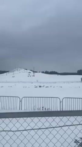 The video shows a snowy hillside with a few people sledding down. The sky is overcast. In the foreground, there's a metal fence, slightly out of focus. Beyond the fence, the hillside is covered in snow. There are a few sparse trees near the top of the hill. Several tall lampposts are scattered across the landscape. Some of the people are at the top of the hill, preparing to sled, while one person is actively sledding down.