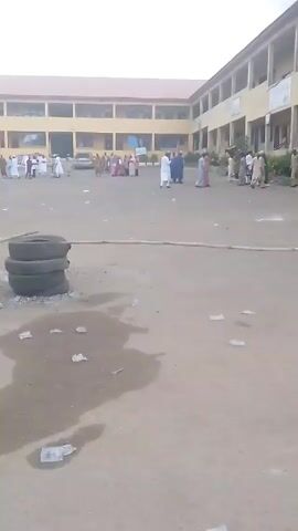 The video depicts a large outdoor area in front of a building, likely a school or public facility. The ground is paved, but covered with litter and damp patches. People are gathered in groups in the background, many dressed in white or light-colored traditional clothing. In the foreground, a young boy wearing a traditional brown tunic and a patterned hat stands looking to the side. There are tires piled in the scene and a horizontal pole laying on the ground. Several cars are parked along one side of the area. The sky is overcast and the overall lighting appears dim.