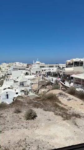 The video shows a panoramic view of a white-washed village perched on a hillside overlooking the sea. The architecture is characterized by cubist buildings with blue-domed churches, typical of the Cycladic islands in Greece. The sky is a clear, bright blue. The village is densely populated with buildings constructed on varying levels of the terrain. Some buildings appear to be hotels or residences with balconies and terraces. There are numerous people walking along narrow pathways and stairs that wind through the village. Some of these pathways are lined with shops, indicated by awnings and displays. The foreground of the video consists of dry, rocky terrain with sparse vegetation, suggesting a Mediterranean climate. The overall impression is a vibrant, sun-drenched tourist destination.